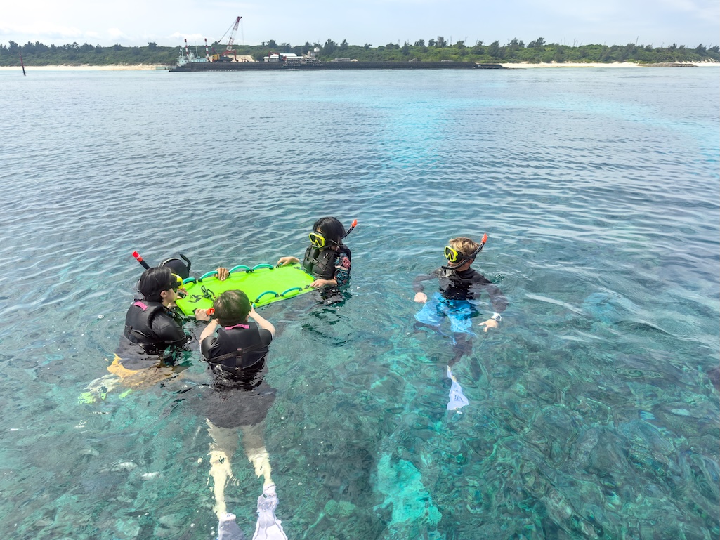 ガイドの指示でフロートボードを掴みながら浅瀬の珊瑚礁を観察する水納島のシュノーケルグループ