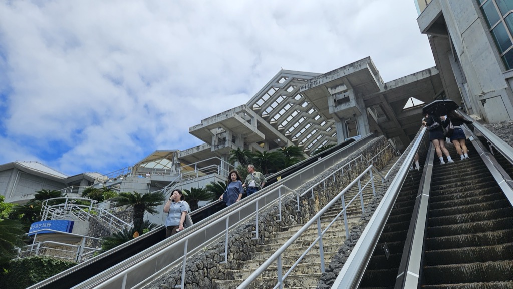 美ら海水族館へと続く長いエスカレーターと階段 特徴的な三角屋根の建築と本部半島の空模様