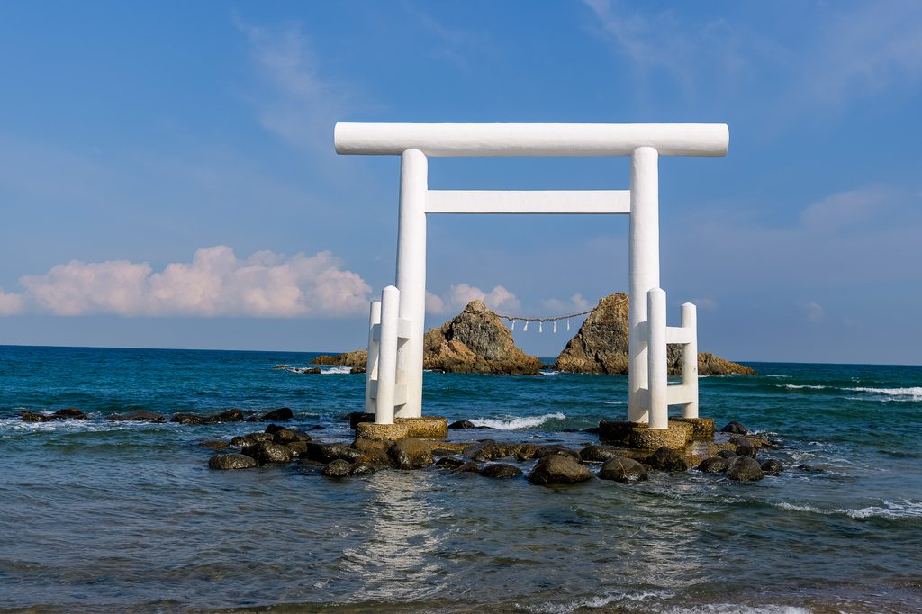 夕陽が夫婦岩の間に沈む瞬間の絶景。空はオレンジ〜紫のグラデーション、海面に光の道が伸びる情景